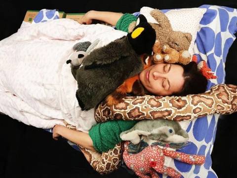10 Lieder - sonst geh ich nicht ins Bett – Person liegt mit geschlossenen Augen auf gemusterten Kissen, umgeben von mehreren Stofftieren, darunter ein Pinguin, Affe und Elefant.