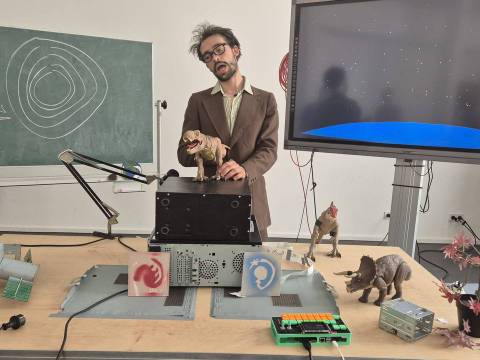  – Person im Anzug mit Spielzeugdinosaurier in der Hand steht vor Tisch mit Computerteilen und Dinosauriern. Tafel mit Kreisen und Text: 15t, 250m, 3s, = 10s.