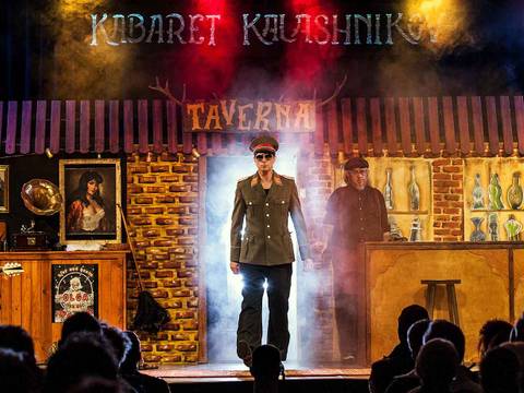 Kabaret Kalashnikov – Bühne mit drei Personen vor einer gemauerten Kulisse. In der Mitte eine Person in Militäruniform, links ein Gitarrist, rechts eine Person hinter einer Bar.