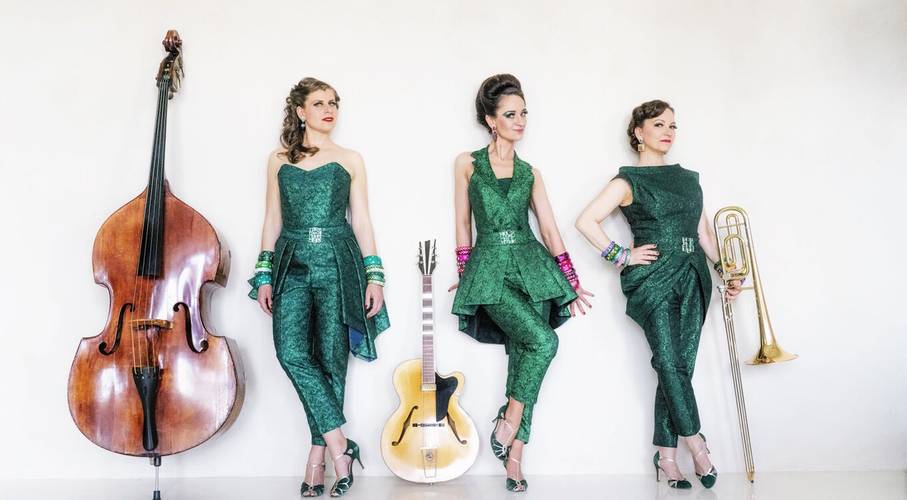 Zucchini Sistaz „Eleganz oder gar nicht!“ – Drei Personen in gruenen Anzuegen, bei einem Kontrabass, einer Gitarre und einer Posaune stehend, tragen Schmuck am Arm und grüne hohe Schuhe.
