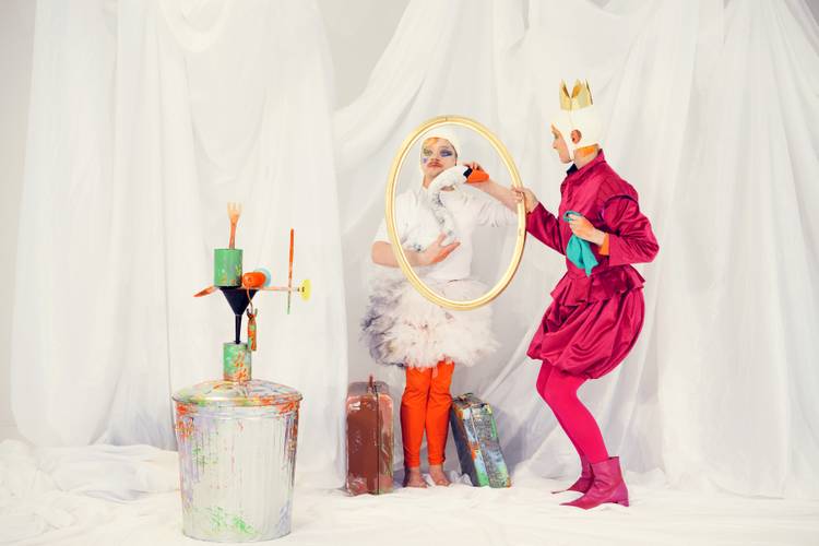 ABC – Das Maler-Alphabet – Person in weißem Tutu und orangefarbenen Strumpfhosen hält goldenen Rahmen. Zweite Person in pinkem Kostüm hält Rahmen fest. Links bemalter Mülleimer.