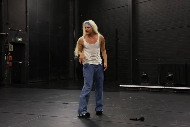 T-Boy on Grindr – Person mit langem blondem Haar und blauem Stirnband trägt weißes Tanktop und blaue Jeans, steht auf leerer Bühne. Mikrofon liegt auf dem Boden.
