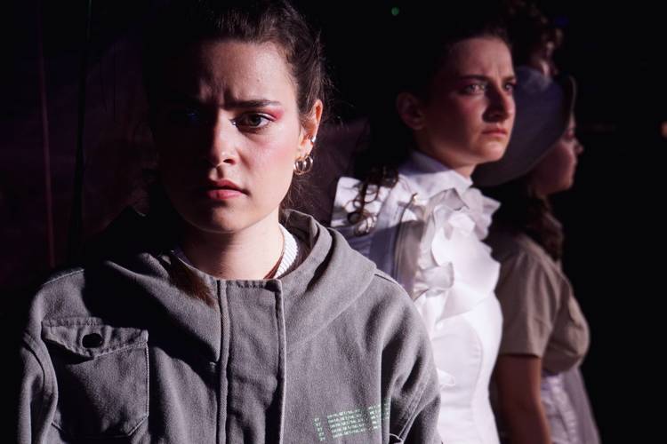 Drei Personen stehen hintereinander. Die vorderste trägt eine graue Jacke, schaut nachdenklich. Die mittlere Person trägt ein weißes Oberteil mit Rüschen.