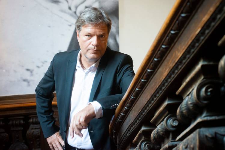 Robert Habeck im Berliner Ensemble (2025) – Eine Person im Anzug lehnt an einem Holzgeländer, schaut ernst. Im Hintergrund ein unscharfes, schwarz-weißes Bild.
