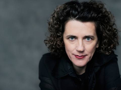 Olga Neuwirth – Person mit lockigem Haar und schwarzer Kleidung blickt direkt in die Kamera vor grauem Hintergrund.