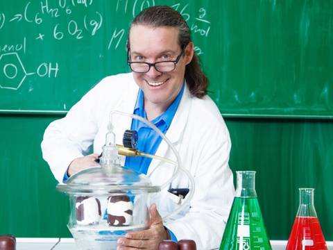CheMagie Show – Person mit Brille und Laborkittel hinter einer Glasglocke. Hintergrund Tafel mit chemischen Formeln, darunter 6 H2O und Phenol.