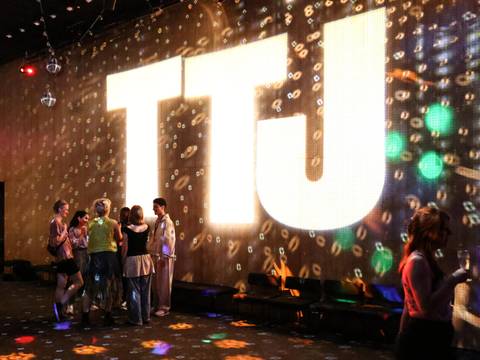 Theatertreffen der Jugend – In einem abgedunkelten Raum wird ein großes TTJ-Logo an die Wand und Lichter an die Wand projeziert. Davor stehen junge Leute in Gruppen.