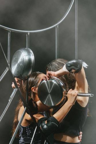 BANG! – Zwei Personen mit Pfannen in den Haenden und Knieschonern stehen dicht beieinander vor einem metallischen Geruest.