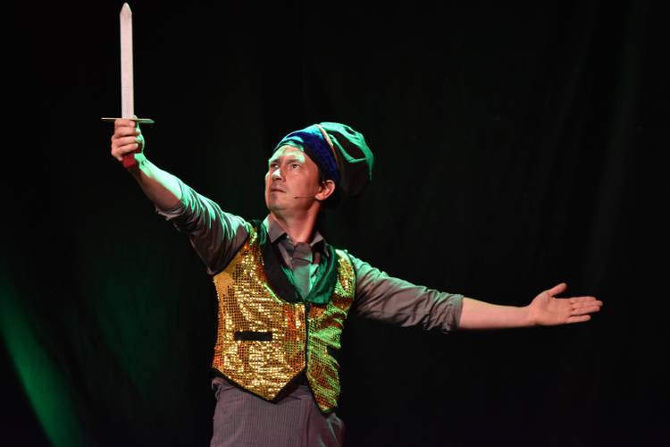 Eine Person in glitzernder Weste und Turban haelt ein Schwert hoch und breitet den anderen Arm aus. Hintergrund dunkel.