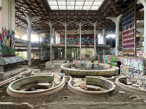  – Verlassener Raum mit verzierten Säulen, bunten Graffiti an Wänden und Decke, mehrere leere Brunnen. Boden voller Schutt und Abfall.