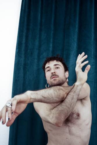 Oberkörperporträt einer shirtlosen Person mit verschränkten Armen in einer tänzerischen Pose vor einem dunkeltürkisen Vorhang; sie trägt Ohrringe und Ringe und blickt direkt in die Kamera.