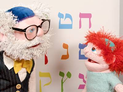  – Zwei Puppen stehen nebeneinander. Die linke Puppe stellt einen jüdischen Rabbi dar. Er trägt eine große Brille mit schwarzem Rahmen, auf dem Kopf trägt er eine Kippa. Rechts neben ihm steht eine Puppe, die ein Kind darstellt. Er ist etwas kleiner als der Rabbi und trägt ebenfalls eine Kippa.