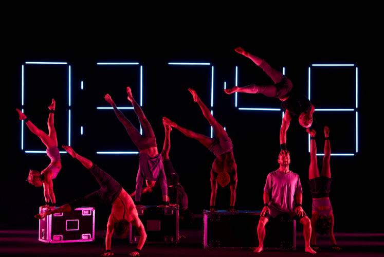 Ten Thousand Hours – 7 Artists machen einen Handstand auf verschiedenen Gegenständen und neigen ihre geöffneten Beine nach links. Ein Künstler sitzt und trägt einen anderen auf seinem Kopf.