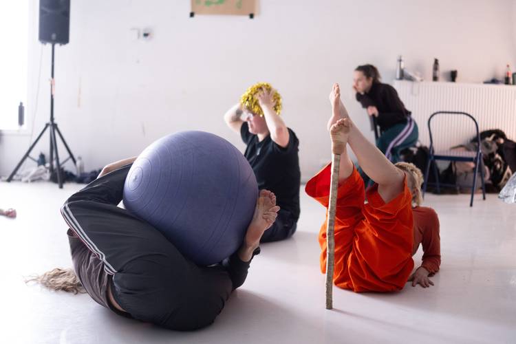 Drei Personen auf dem Boden in verschiedenen Posen, eine mit einem Gymnastikball, eine mit einem Stock, eine trägt Glitzer auf dem Kopf.
