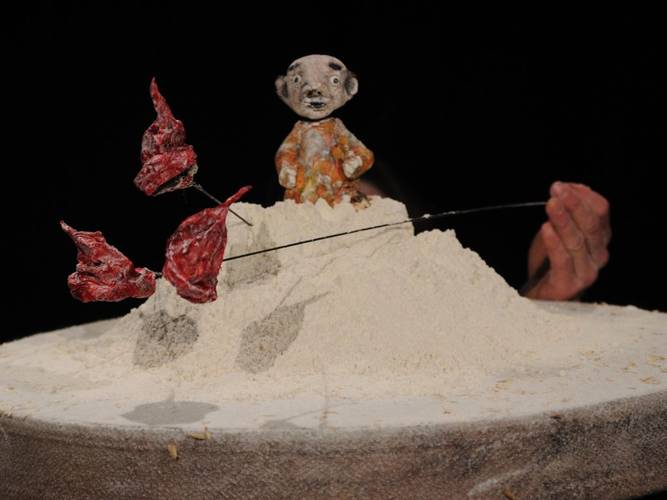 Tonfigur auf Sandhügel, links rote Blätter an Draht, rechts Hand, schwarzer Hintergrund.
