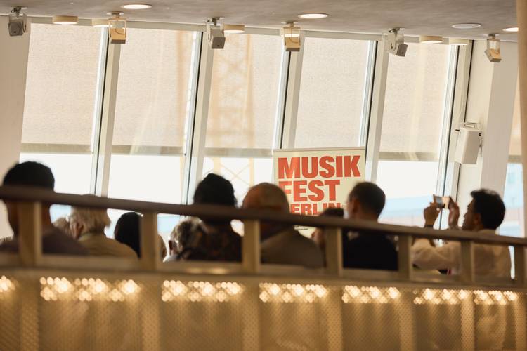 Personen vor einem Fenster; Plakat mit rotem Text "MUSIK FEST BERLIN" im Hintergrund; männlich gelesene Person macht ein Foto.