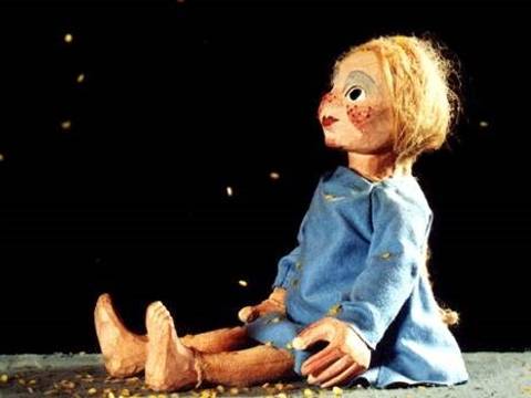 Sterntaler – Holzmarionette mit blauem Kleid und geflochtenem Haar sitzt auf grauem Boden; goldenes Konfetti in der Luft schwebend.