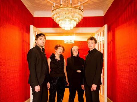  – Vier Personen in schwarzer Abendgarderobe stehen in einem elegant beleuchteten Flur mit leuchtend roten Wänden, weißen Türrahmen und zwei großen Kristalllüstern. Die Gruppe blickt in die Kamera und ist mittig im Bild positioniert. Die warme Beleuchtung und die symmetrische Perspektive verleihen der Szene eine festliche, stilvolle Atmosphäre.