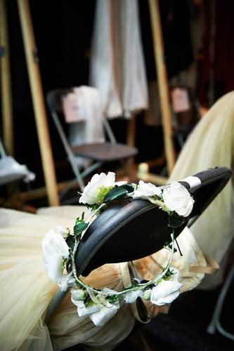 Backstage-Führung – Blumenkranz aus weißen Rosen liegt auf einem Stuhl. Im Hintergrund unscharfe Kleider auf Kleiderbügeln und ein weiterer Stuhl.