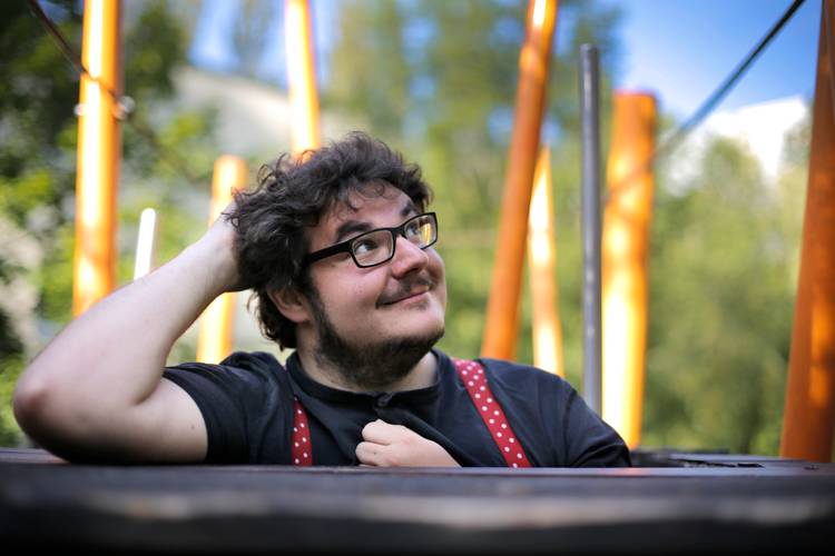 © Dennis Pauls – Mann mit Brille, der sich auf einen Tisch stützt und in die Ferne schaut, mit großen orangefarbenen Stangen im Hintergrund.