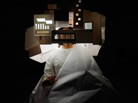 GPO Box No. 211 – Eine Person in weißem Gewand hält einen großen Papierbogen, Kopf von geometrischen Formen umgeben, auf schwarzem Hintergrund.