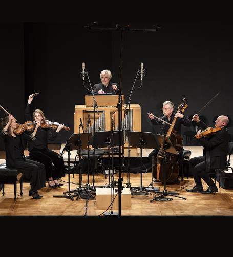 Eine Person spielt ein Harmonium, umgeben von vier Musiker*innen mit Streichinstrumenten auf einer Bühne mit Mikrofonen davor.