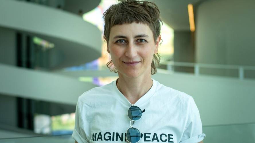 Ulrike Almut Sandig – Person mit Kurzhaarschnitt, hellem T-Shirt mit Text "IMAGINE PEACE", Sonnenbrille hängt am Kragen. Hintergrund mit gewundenem, innenarchitektonischem Detail.