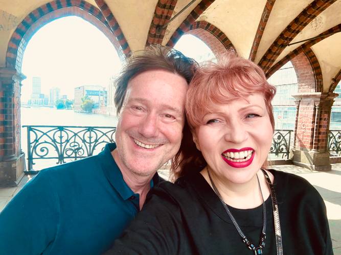 Rainer Biefeldt und Bettina Meske – Rainer Bielfeldt und Bettina Meske, beide lächelnd, machen ein Selfie unter einer historischen Backsteinarkade an der Berliner Oberbaumbrücke mit Blick auf die Spree.
