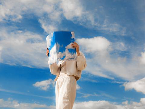  – Person in hellem Outfit haelt ein großes Foto eines Himmels mit Wolken vor dem Gesicht, das sich nahtlos in den echten blauen Himmel mit Wolken einfügt.