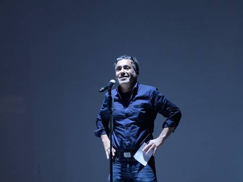 Jürgen Kuttner, Foto: Thomas Aurin – Person steht mit Mikrofon auf Bühne, trägt dunkles Hemd und Jeans, Papier in der linken Hand, neutrale Mimik, dunkler Hintergrund.