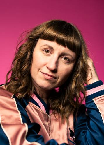 Paula Fürstenberg – Person mit braunen Locken, Ponyfrisur und rosa-blauer Jacke vor pinkem Hintergrund, schaut in die Kamera und fasst mit einer Hand an die Haare.
