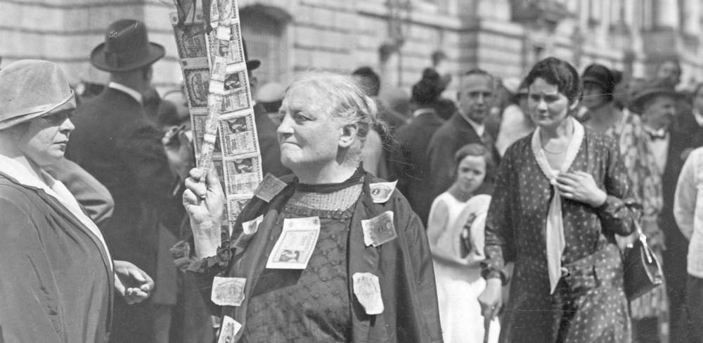 Fotografie „Eine Rentnerin protestiert mit aufgenähten Banknoten gegen die Geldentwertung“ – Schwarzweißfoto mit älterer Person, die ein Schild mit Bildern und Text haelt. Kleidung mit Geldscheinen. Menschenmenge im Hintergrund.