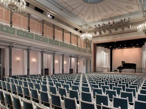  – Großer Konzertsaal mit Reihen blau gepolsterter Stühle, Kronleuchter, Bühne mit schwarzem Flügel, rosa Wände und einem dekorativen Deckenmuster.