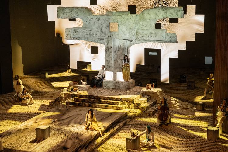 Mittwoch aus Licht – Bühnenbild mit großem steinernen Kreuz, umgeben von Personen und Musikinstrumenten in einer wüstenartigen Landschaft. Eine Person steht zentral.