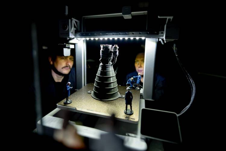 Transport: Fasten your Seatbelts! – Miniatur-Weltraumantrieb unter hellem Licht; zwei Unschärfen männlich gelesener Personen im Hintergrund; vier Miniaturfiguren um das Gerät gruppiert.