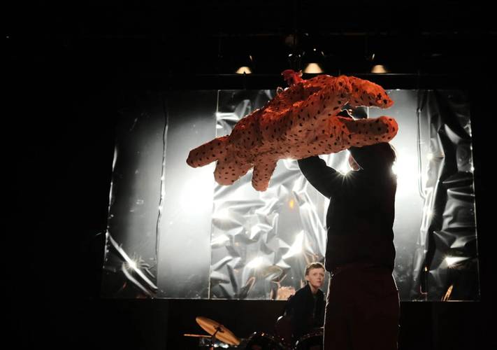Person hebt eine große, orange gepunktete Leopardenmarionette in die Luft. Hintergrund mit reflektierendem Material und Schlagzeug.