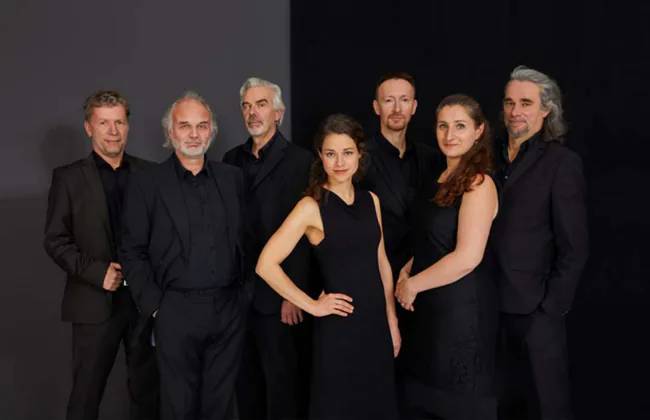L'ART DE PASSAGE - Vom Tango zum Jazz und zurück – Sieben Personen in schwarzer Kleidung stehen vor dunklem Hintergrund, sechs männlich gelesene und eine weiblich gelesene Person in der Mitte, alle schauen zur
