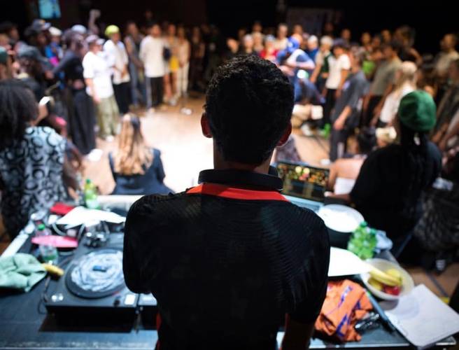 Person am DJ-Pult vor einer Menschenmenge auf einer Veranstaltung, von hinten gesehen. Laptop und Turntables vor der Person.