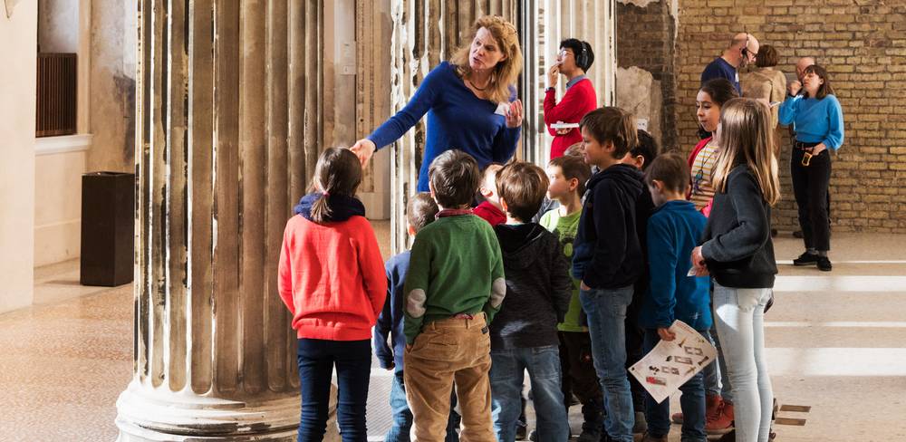 Familienführung im Neuen Museum – Ein*e Erwachsene in blauem Pullover spricht mit einer Gruppe von Kindern in einem Raum mit antiken Saeulen und Ziegelmauerwerk.