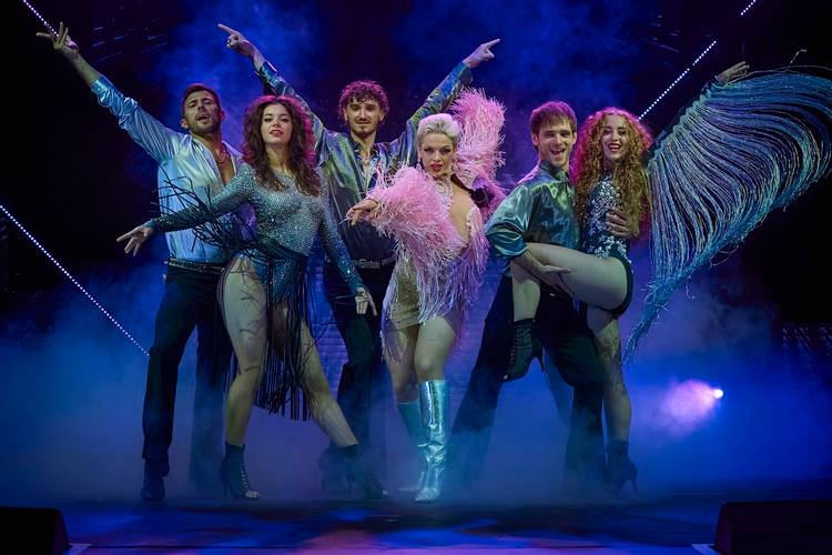Disco Dancers – Sechs Tänzer*innen in Posen, bunte Bühnenkleidung mit Fransen, blauer und violetter Nebel, strahlende Lichter im Hintergrund.