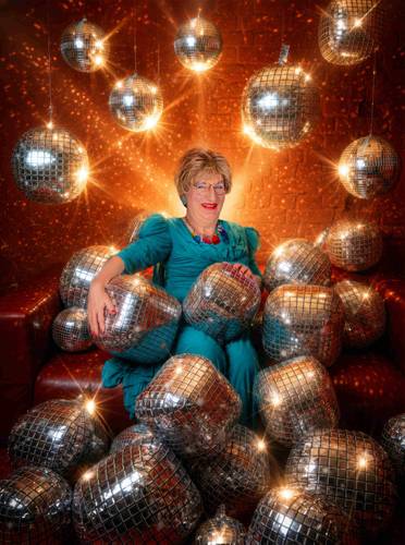 Edith Schröder – Person in blauem Kleid sitzt auf rotem Sofa, umgeben von glitzernden Discokugeln, die von der Decke hängen und auf dem Boden liegen.