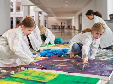 Familienworkshop in der Neuen Nationalgalerie – Kinder in weißen Kitteln malen mit Pinseln auf großen Leinwänden auf dem Boden in einer hellen Halle. Farbtöpfe sind um sie herum verteilt.