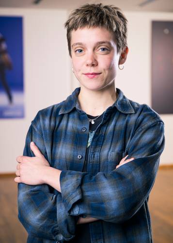 Person mit kurzem Haar und Ohrring, tragend kariertes Hemd mit verschränkten Armen, steht vor unscharfer, gerahmter Kunst.
