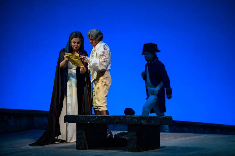 Tosca – Zwei Personen in historischer Kleidung lesen ein Dokument, blaue Bühnenbeleuchtung, Figur im Hintergrund trägt Hut und Mantel, Steinbank im Vordergrund.