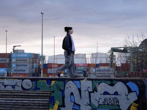 © Julia Krewani und Ralph Etter – Eine Person läuft in beginnender Dunkelheit auf einer Mauerkante, im Hintergrund Container.
