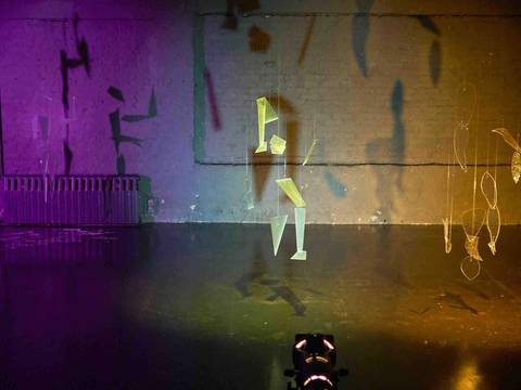  – Objekte aus Draht und durchsichtigen Materialien hängen vor einer bunt beleuchteten Wand. Schattenspiele und Lichtprojektionen sind sichtbar.