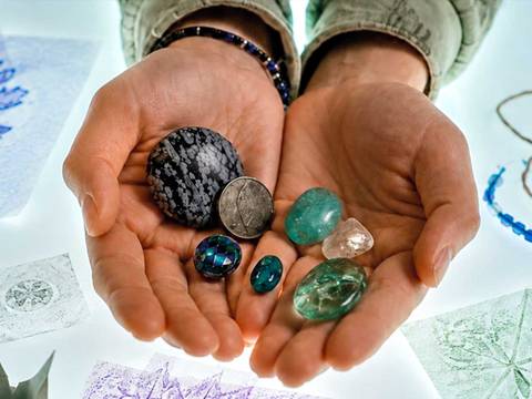 Workshop: Unbezahlbar – Zwei Hände halten verschiedene glatte Steine und eine Münze auf hellem Hintergrund mit Blättermustern, Faden und blauer Perlenkette.