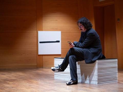  – Person mit Anzug sitzt auf Holzbank, schaut auf Smartphone. Im Hintergrund ein Bild mit schwarzem horizontalem Strich auf weißem Hintergrund. Holzwände.