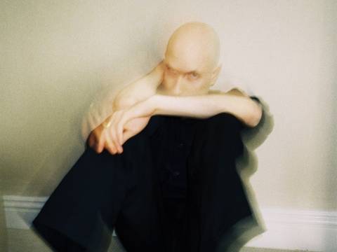 Daniel Blumberg, Foto: Taylor Russell – Glatzköpfige Person in schwarzer Kleidung, sitzt unscharf mit angewinkelten Beinen gegen eine Wand. Hände verschraenken vor dem Gesicht. Schwarze Stiefel.