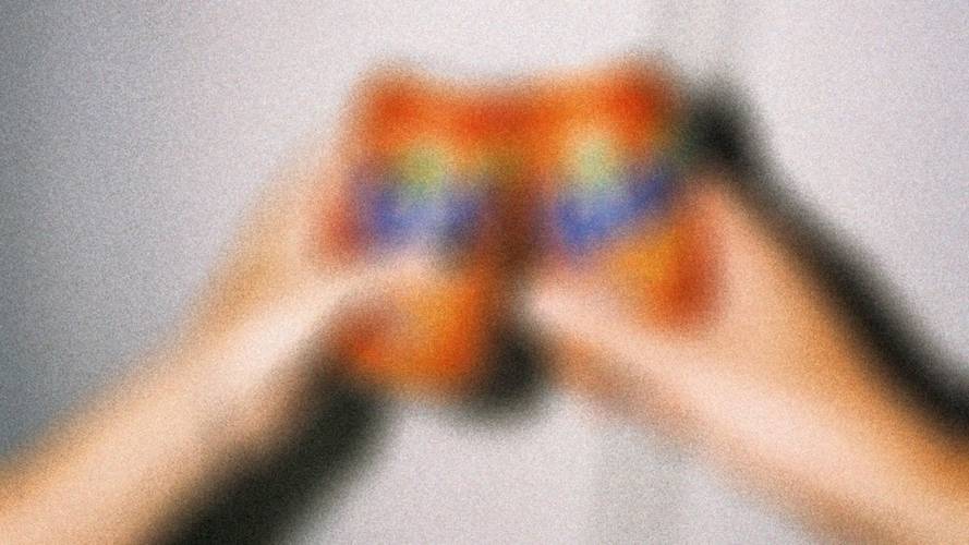 Zwei verschwommene, einander zuprostende Haende halten orangefarbene Getraenkebecher mit Regenbogenmuster. Hintergrund unscharf.